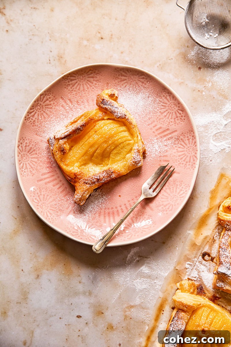 A single pear tart dusted with icing sugar, served on a pink plate with a dessert fork.