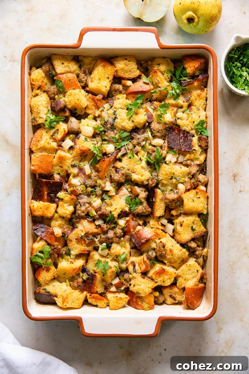 Overhead view of baked sausage stuffing in a rectangular dish, alongside a bowl of diced pear and fresh herbs, showcasing the key ingredients.