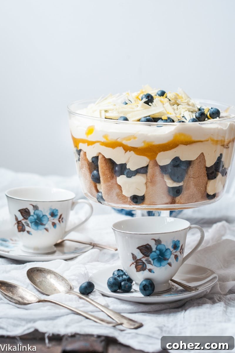 A beautifully layered lemon blueberry trifle with fresh blueberries on top, set on a table with white and blue teacups.