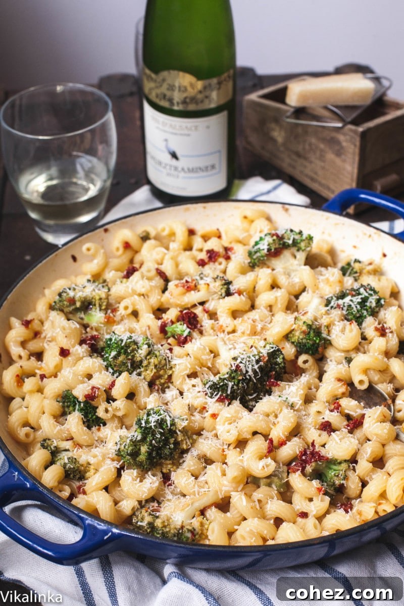 Overhead shot of broccoli pasta in a blue pan, with a bottle of wine, wine glasses, and a block of Parmesan cheese in the soft-focus background