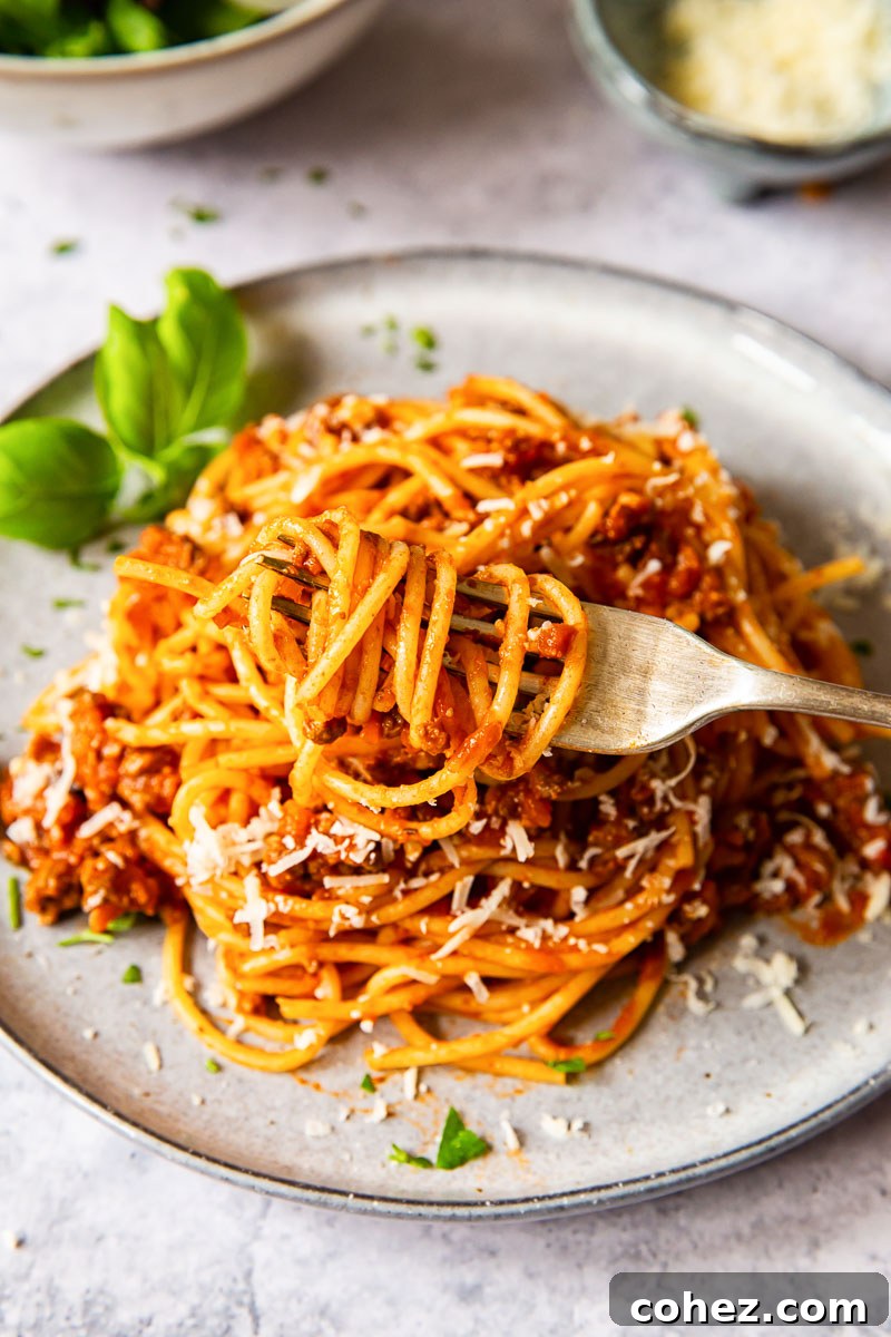 a fork with pasta coiled up with the pasta and meat sauce on a plate behind