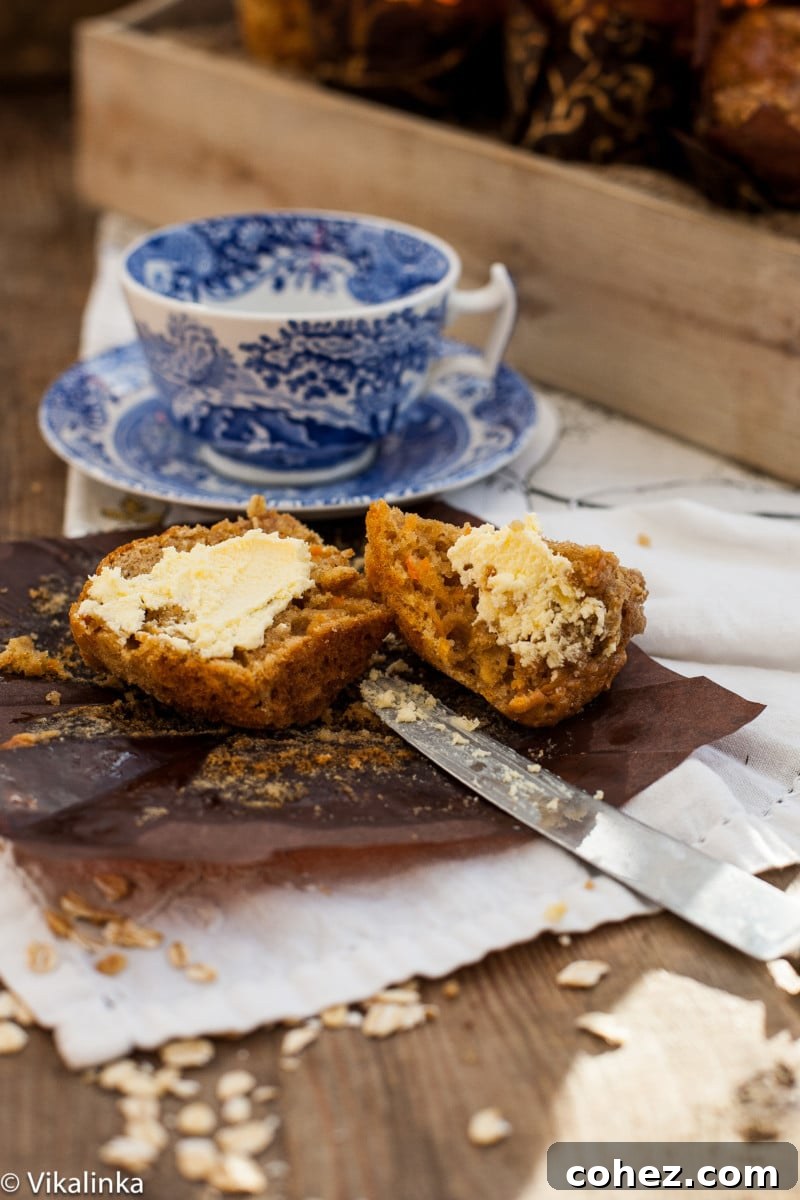 A close-up shot of a carrot oatmeal muffin cut in half, revealing its moist crumb and oat flecks. A blue teacup sits softly blurred in the background, suggesting a cozy snack time.