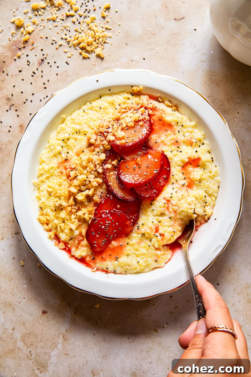 A top-down view of a steaming bowl of millet porridge, with a hand gently reaching in with a spoon, ready to enjoy. The porridge is topped with colorful plums and seeds.