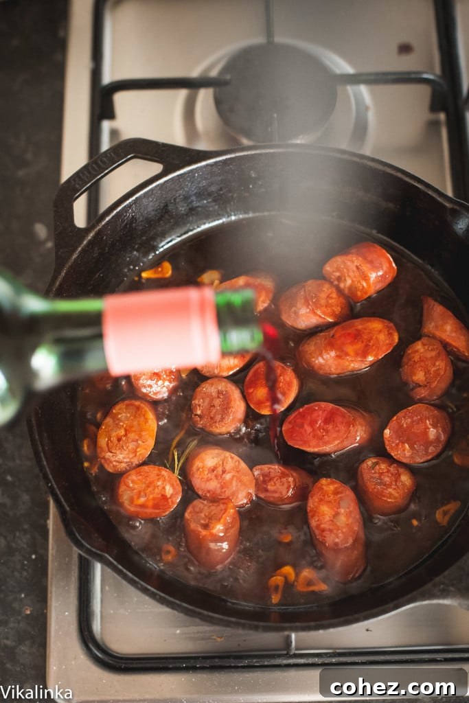 A cook's perspective: Red wine being poured into a hot cast iron skillet containing browned chorizo slices, garlic, and rosemary sprigs, setting the stage for the rich sauce.