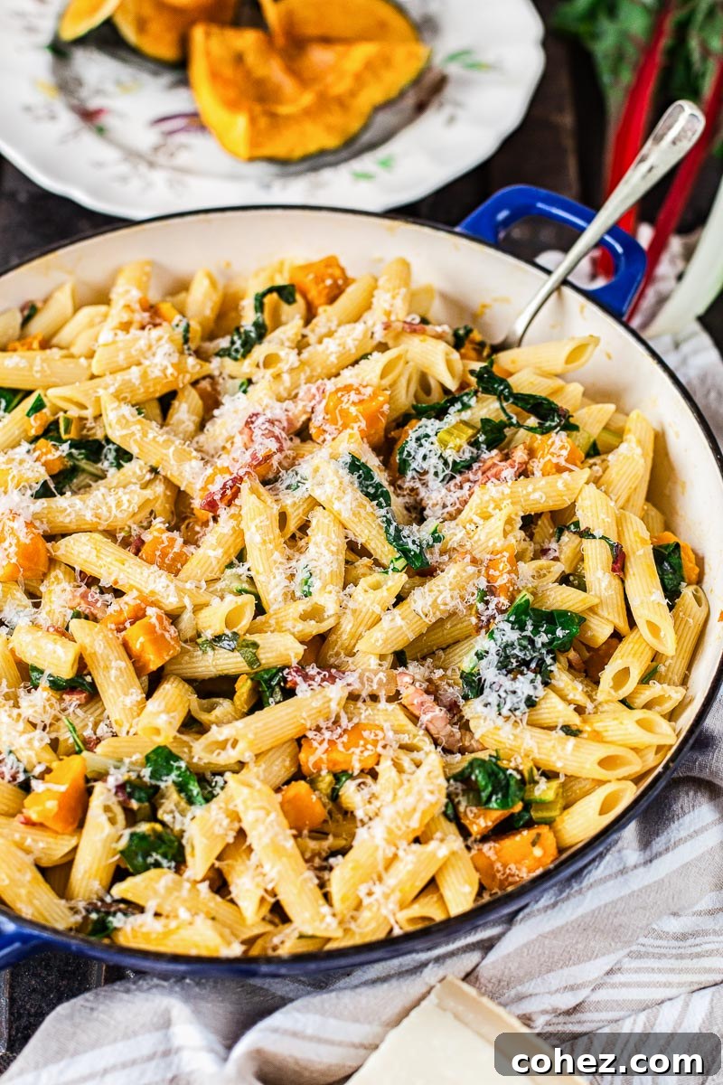Penne pasta expertly tossed with roasted squash and crispy bacon in a large frying pan, with more golden roasted squash pieces visible in the background, ready for final seasoning.
