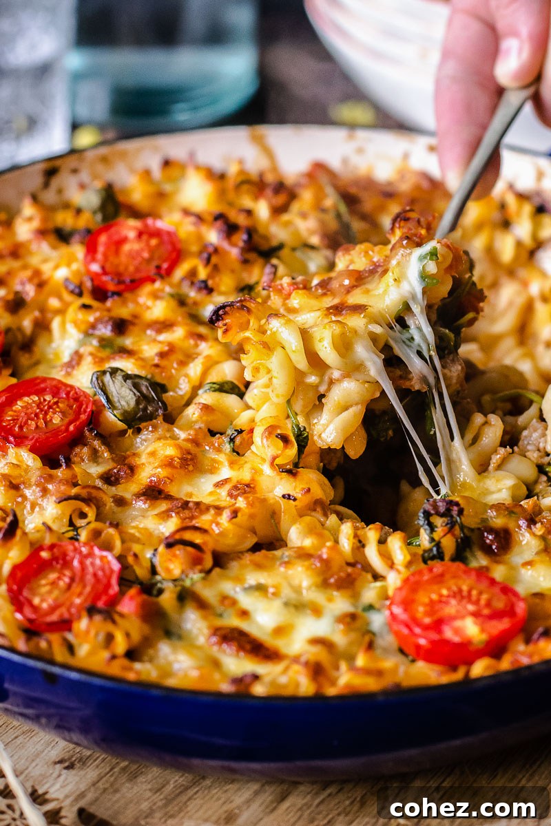 A close-up side view of a rich sausage pasta casserole with a spoon scooping out a generous portion, showing melted cheese and sauce