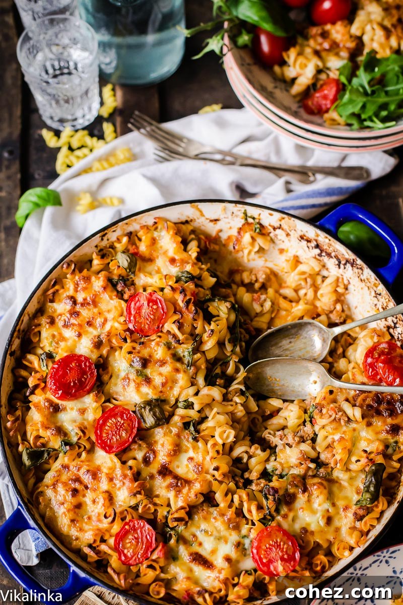 Overhead shot of a bubbling hot sausage pasta bake fresh out of the oven, adorned with glistening melted cheese and bright cherry tomatoes. Background shows serving bowls and glasses, hinting at a family meal.