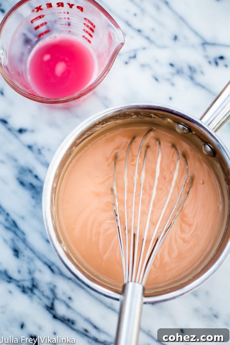 A stainless steel saucepan on a stovetop, containing a bubbling pink rhubarb curd mixture, being actively stirred with a metal whisk to ensure smoothness.