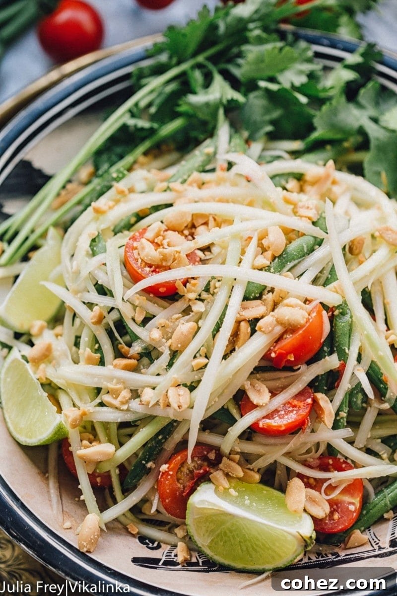 Fiery Som Tam Papaya Salad 6 Close-up of a serving of Thai green papaya salad, highlighting the shredded papaya, vibrant cherry tomatoes, and crunchy peanuts, with a lime wedge.