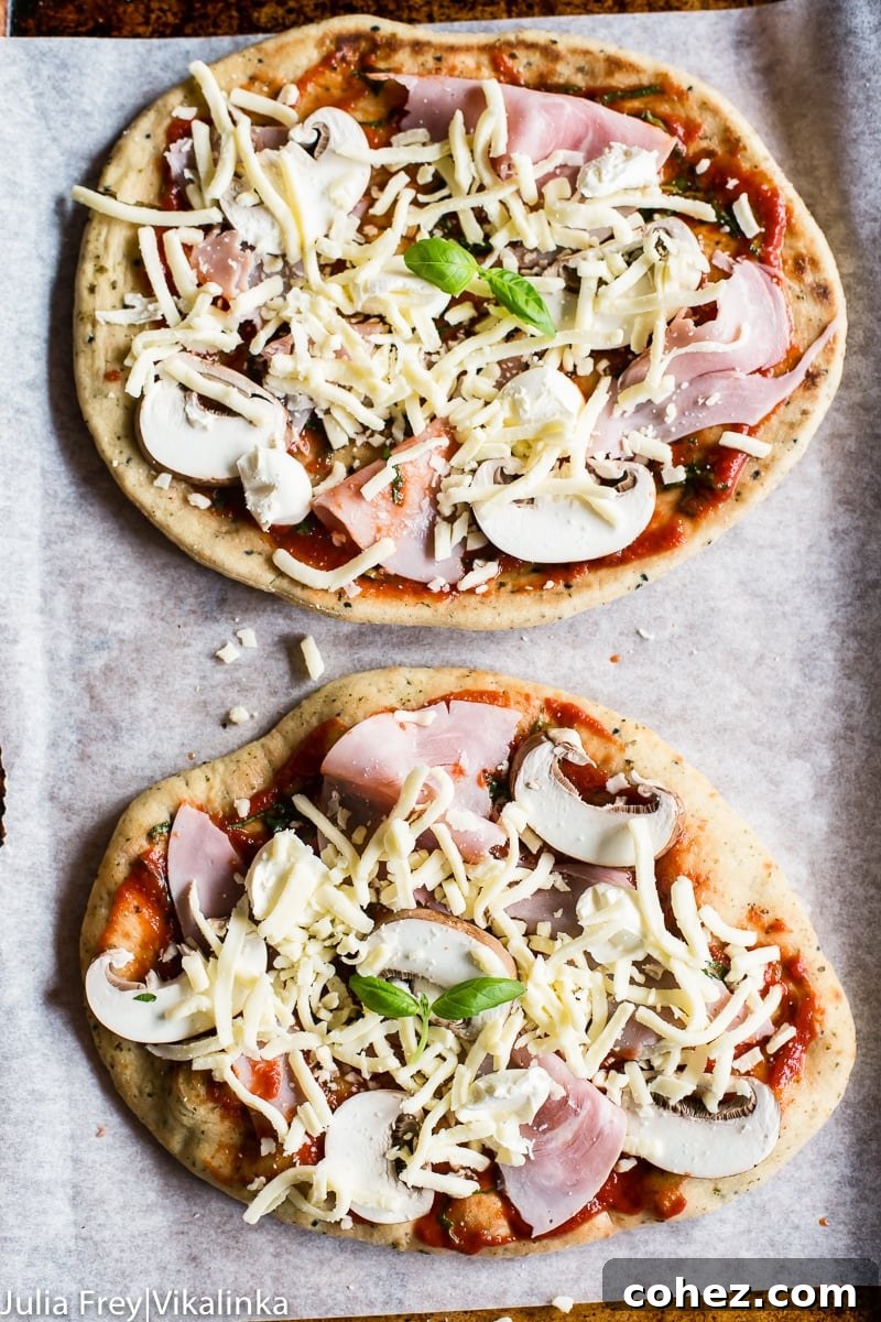 Two unbaked naan pizzas on a baking sheet, topped with sauce, cheese, and vegetables