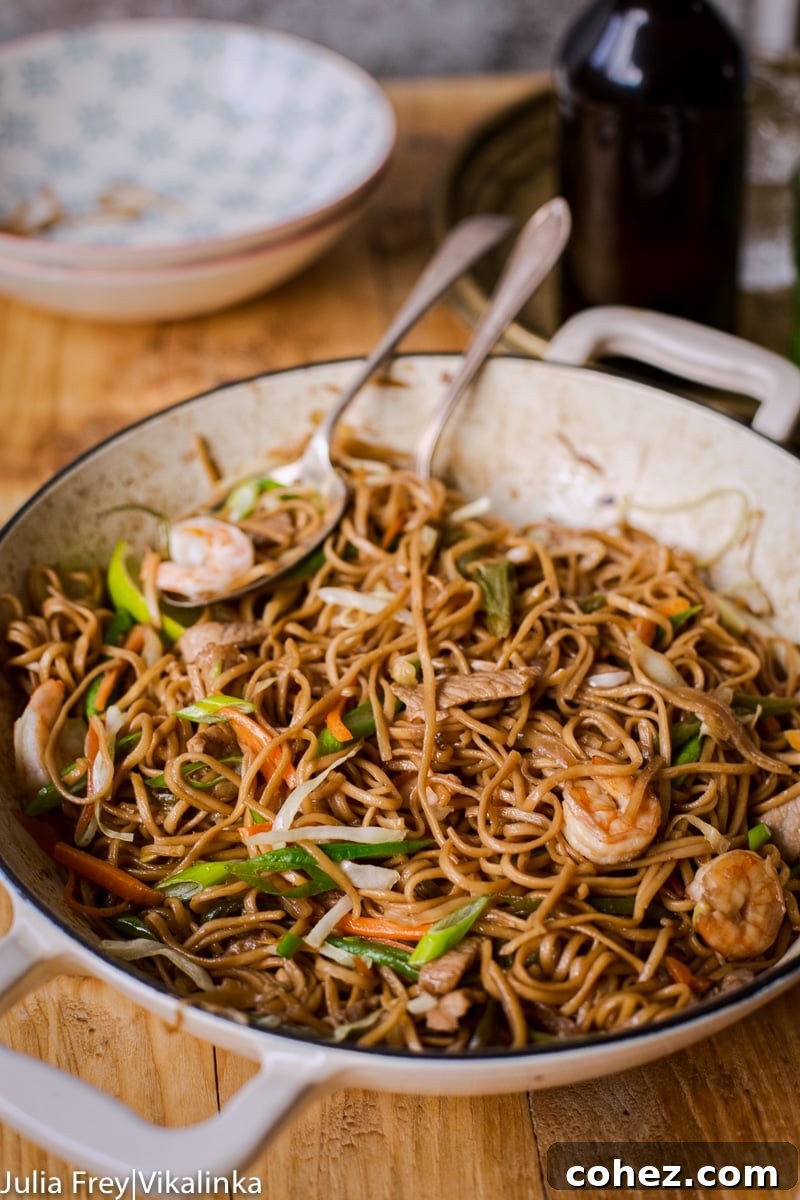 A large pan filled with freshly cooked Pancit Canton, featuring noodles, shrimp, pork, and mixed vegetables, with serving bowls in the background.