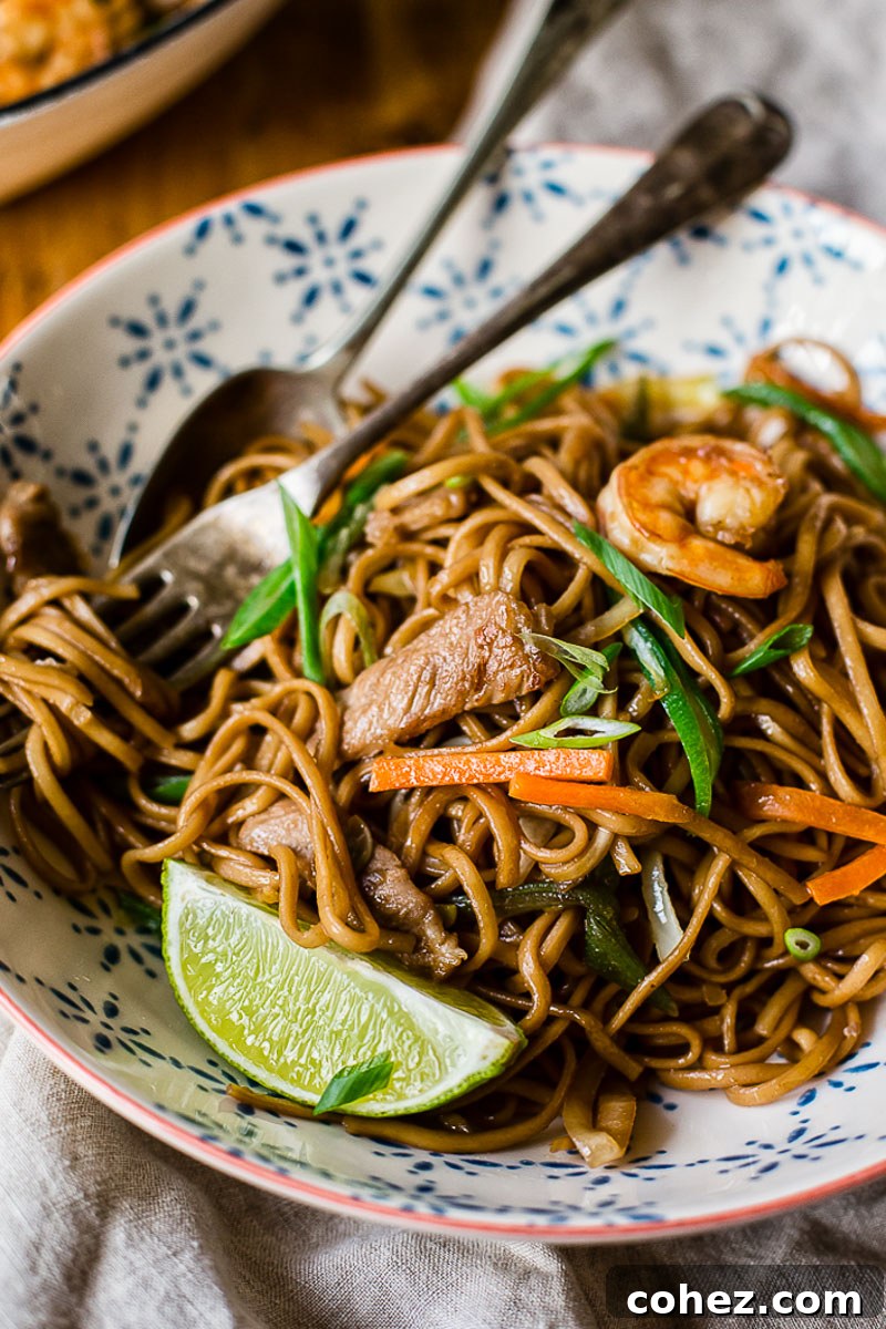 A bowl of delicious Filipino Pancit Noodles with pork and shrimp, garnished with a lime wedge and served with a fork and spoon.