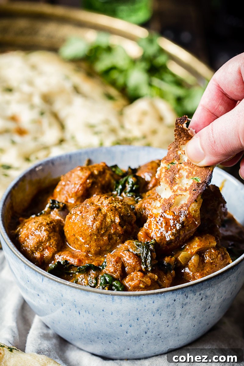 A person dipping a piece of flatbread into a blue bowl of Indian Kofta Curry, showcasing the tender meatballs and rich sauce.