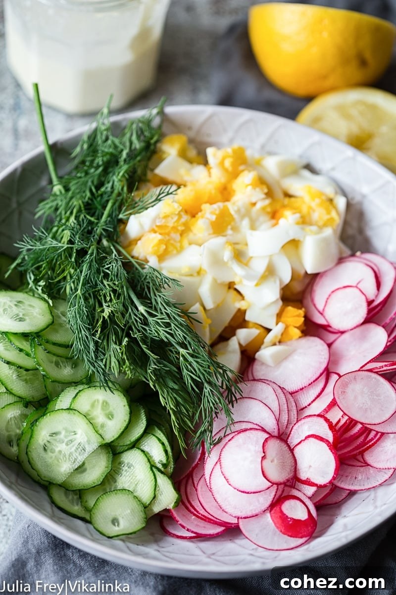 Close-up of thinly sliced cucumbers and radishes, chopped hard-boiled eggs, and fresh dill in a mixing bowl, with a lemon in the background.