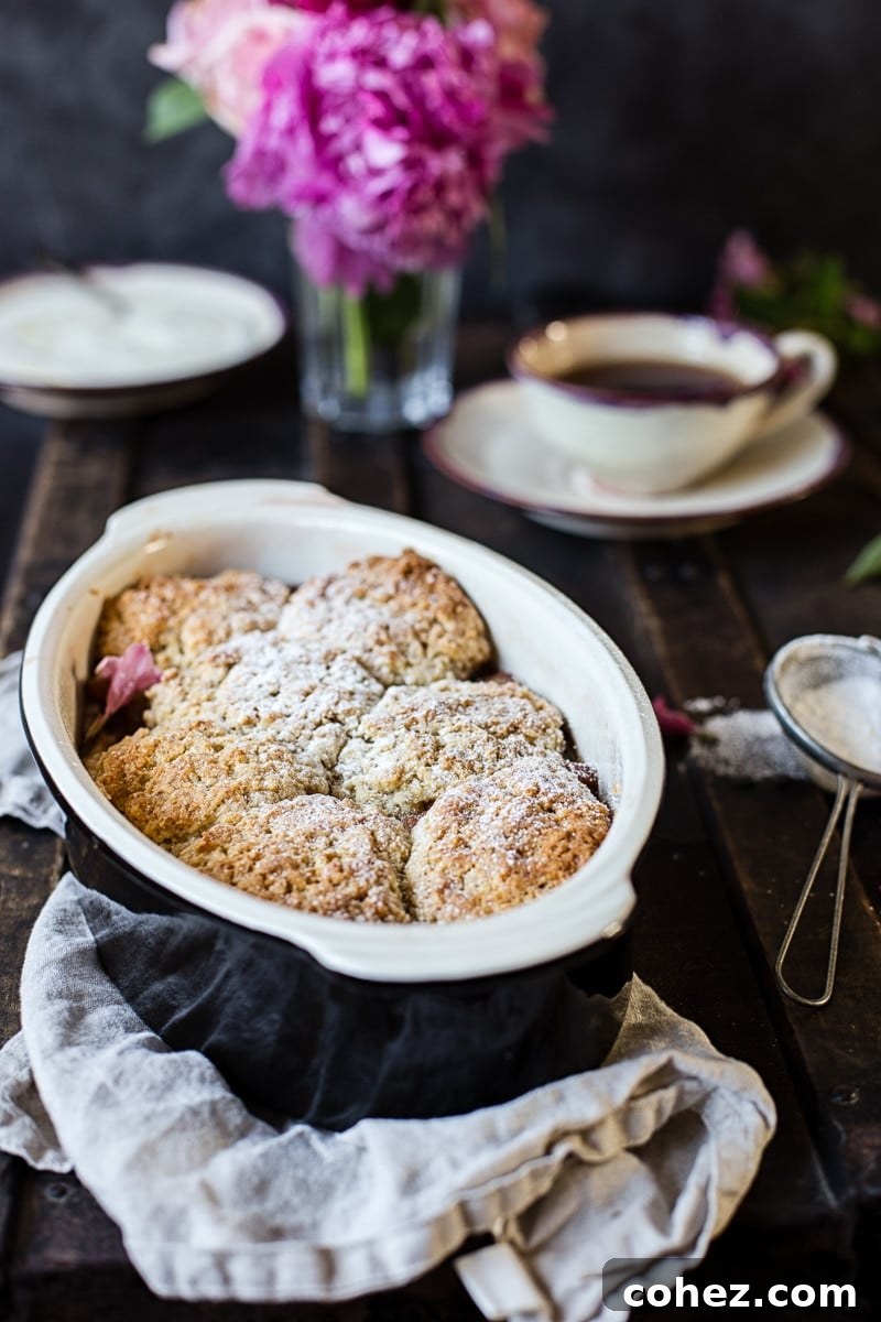 Rhubarb cobbler in a baking dish, freshly baked and steaming, placed in front of a tea setting and fresh flowers.