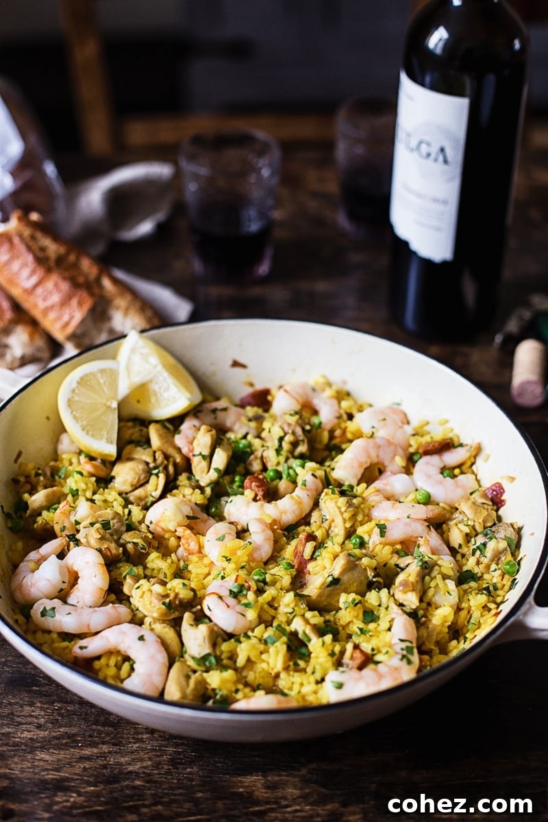 A rustic white pan filled with Spanish rice and shrimp, accompanied by a crusty baguette and a glass of red wine in the background.