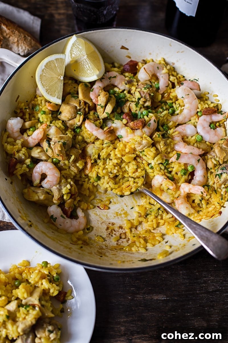 A white pan filled with Spanish rice, chicken, and shrimp, with a serving spoon resting in it. A small white plate of the finished dish sits alongside.