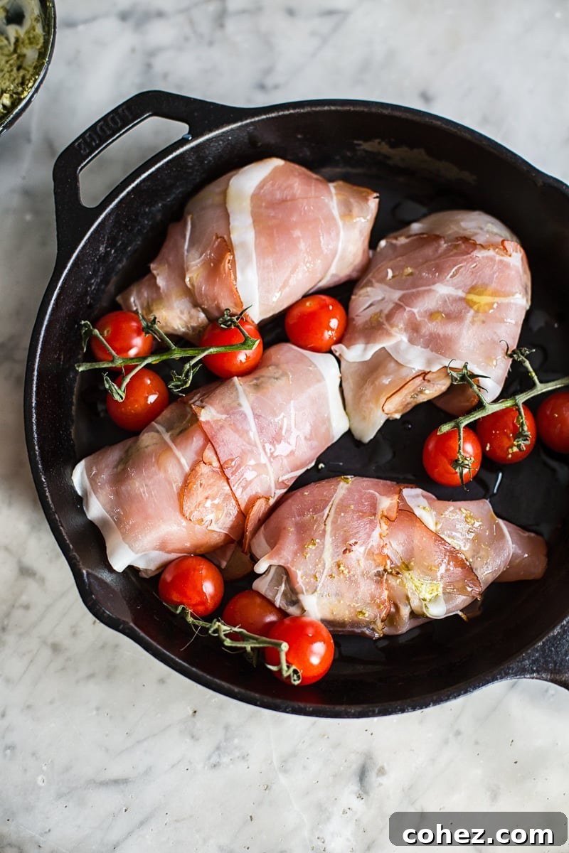 Uncooked prosciutto wrapped pesto chicken parcels on a cast iron pan with cherry tomatoes