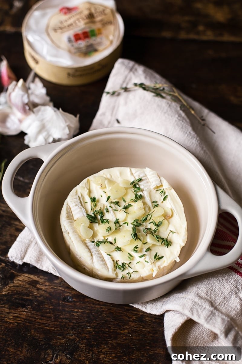 Close-up of a wheel of Brie cheese in a small baking dish, ready for the oven, with garlic slices and thyme sprigs arranged on top.