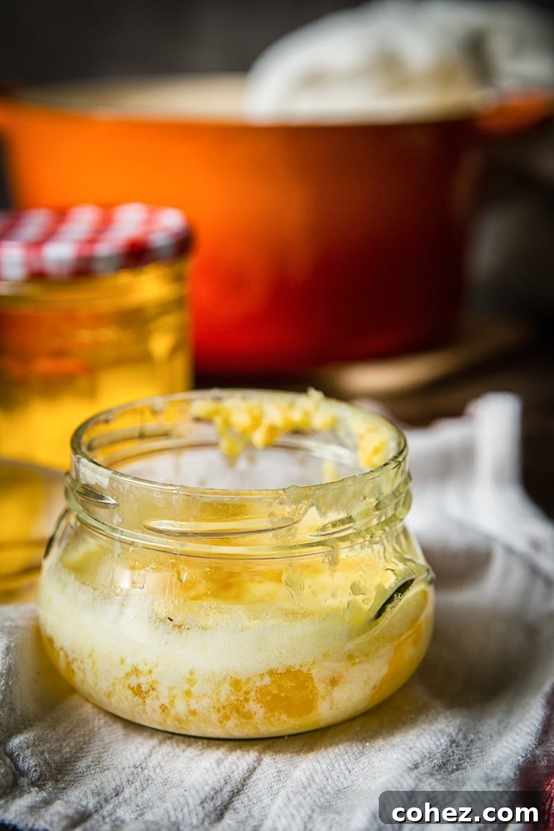 Two jars of golden clarified butter, one with a scoop, sitting beside a small bowl of savory milk solids.