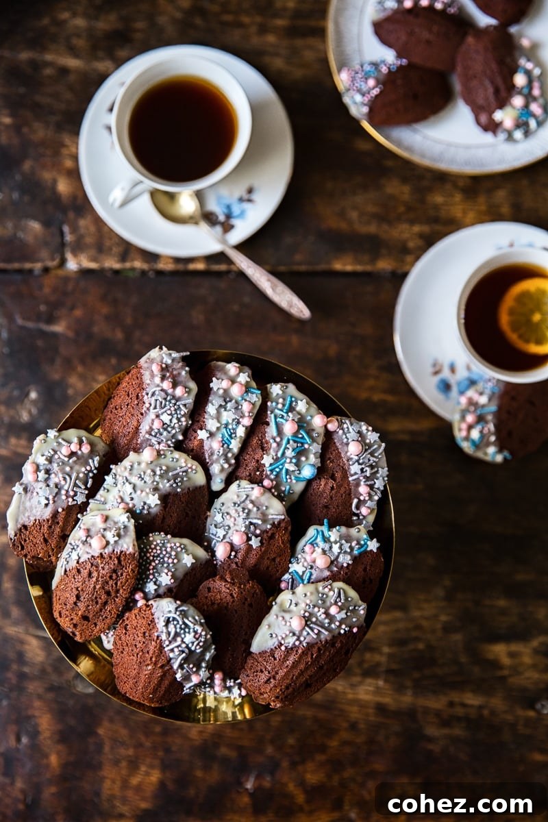 Decadent Chocolate Madeleines 4 An overhead view of a collection of Chocolate Madeleines, some dipped in white chocolate and decorated with unique, colorful sprinkles, next to steaming cups of tea.
