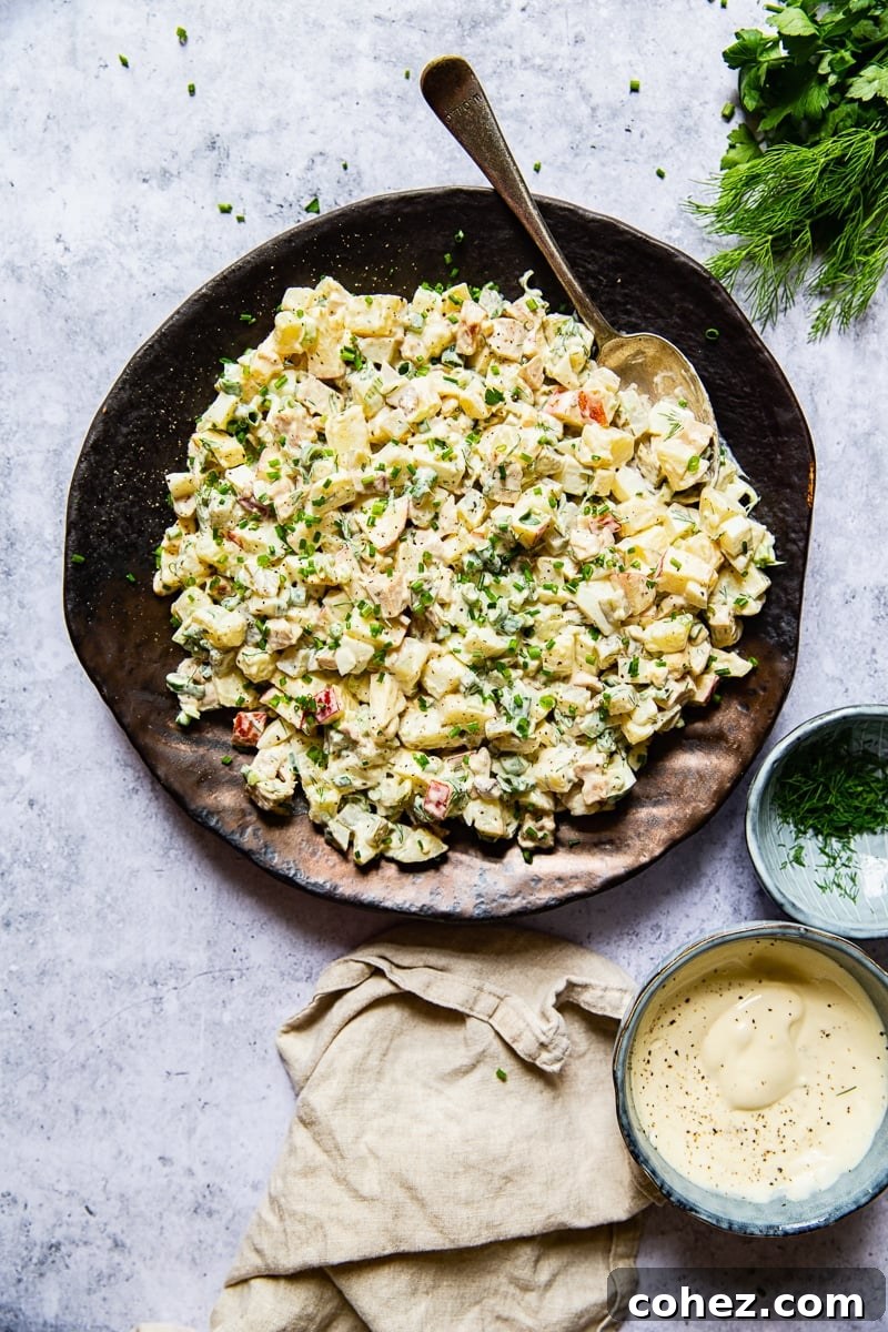 Beautifully presented potato salad on a platter with a serving spoon, accompanied by a cream napkin, a small bowl of salad dressing, and a bowl of fresh dill.