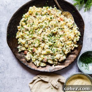 Beautifully presented potato salad on a platter with a serving spoon, accompanied by a cream napkin, a small bowl of salad dressing, and a bowl of fresh dill.