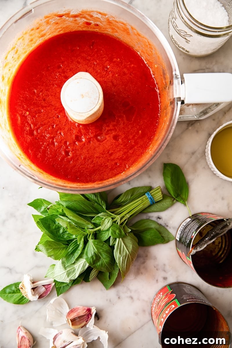 Food processor filled with freshly made red sauce, with basil leaves and empty tomato cans nearby on a kitchen counter