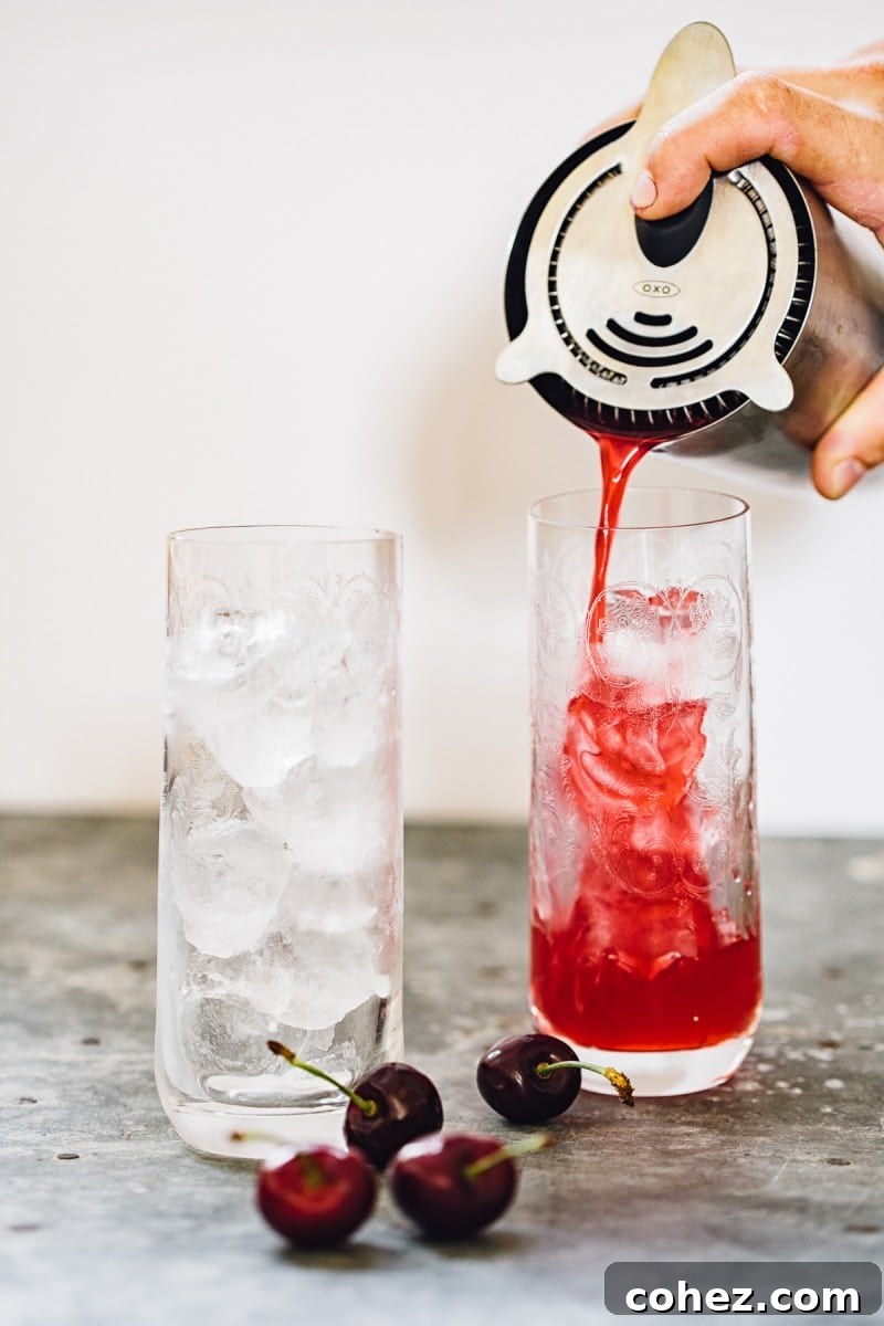 Bartender pouring the cherry Long Island iced tea mixture over ice into elegant glasses with a cocktail shaker in the foreground.