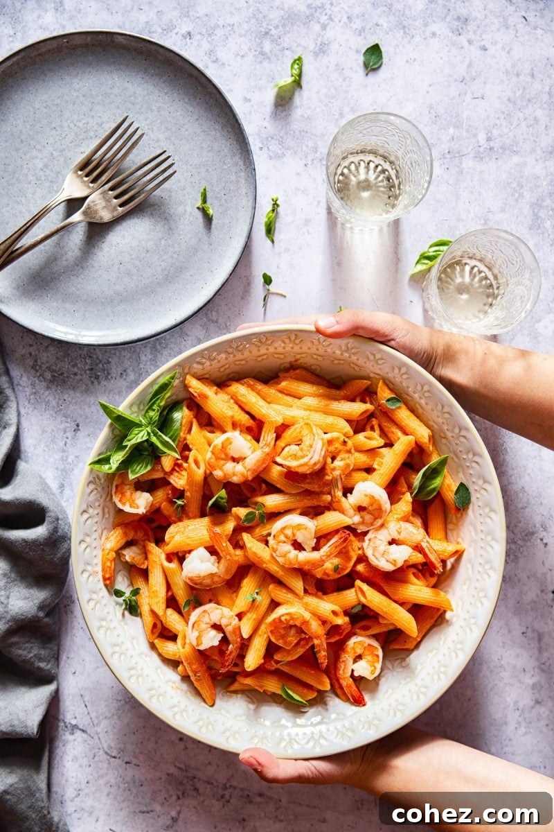 Two hands holding a serving bowl of shrimp penne alla vodka, with glasses and a plate in the background