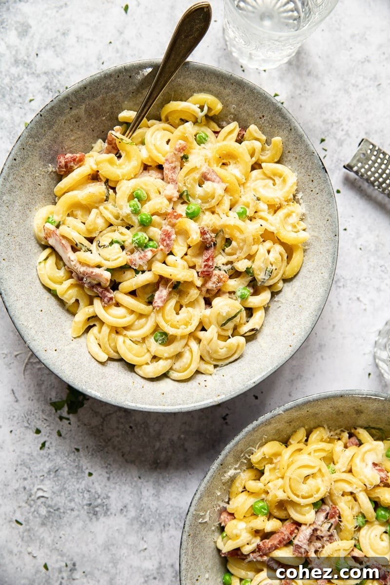 Top-down photo of two bowls of pasta with a fork inserted, highlighting the creamy sauce, pancetta, and peas.