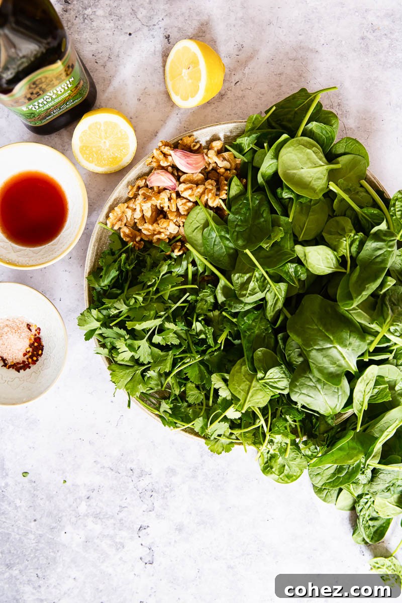 A colorful array of fresh ingredients laid out on a wooden surface, including spinach, cilantro, parsley, walnuts, garlic, lemons, and chili flakes, ready for making spinach pesto.