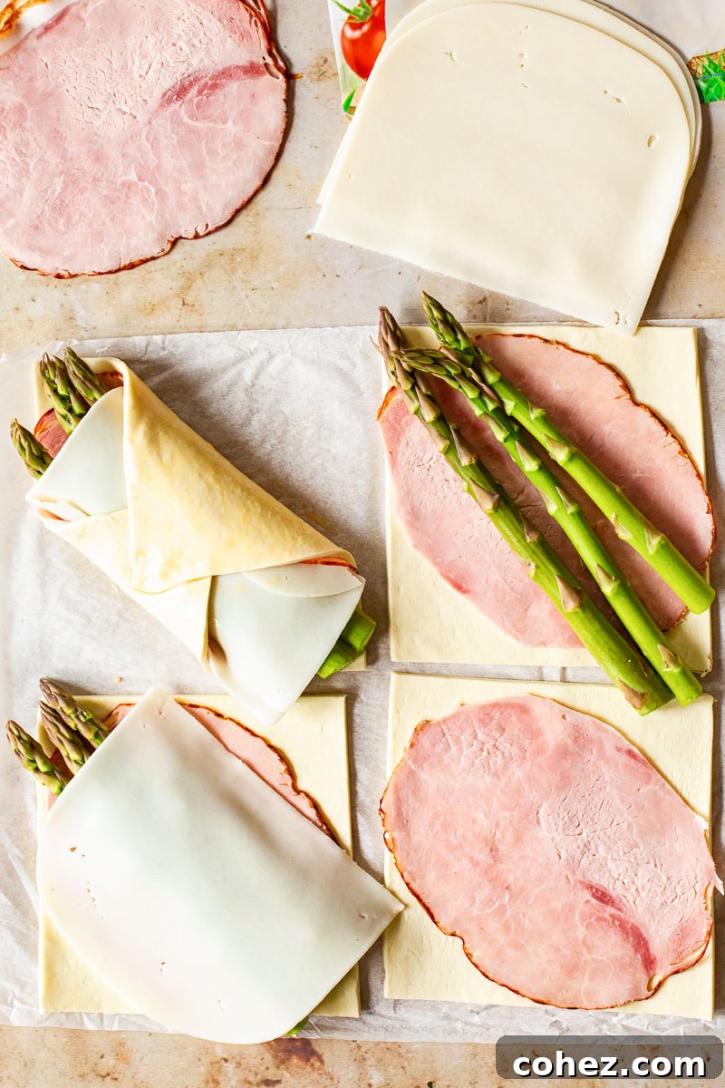 Close-up of unbaked puff pastry squares filled with slices of ham, cheese, and fresh asparagus spears, arranged on a baking sheet.