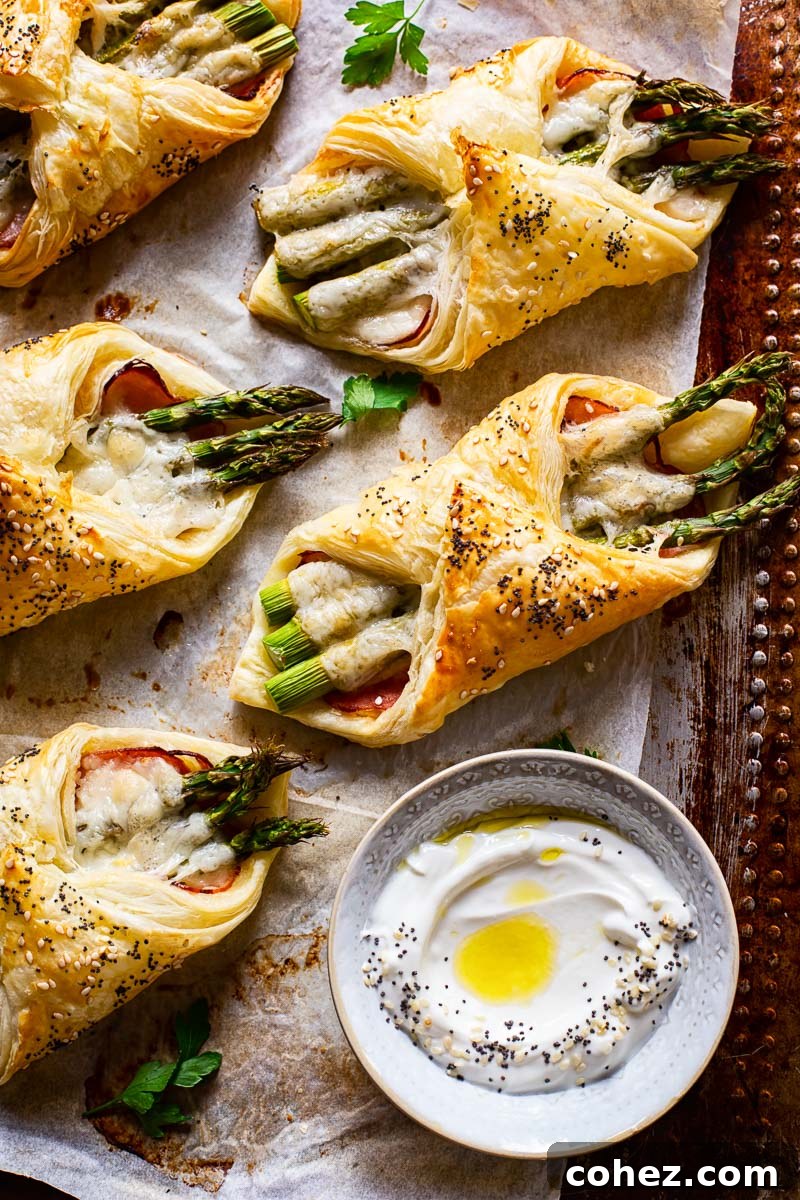 Freshly baked golden cheese and asparagus puff pastry pockets on a parchment-lined baking sheet, with a small dish of dipping sauce nearby.