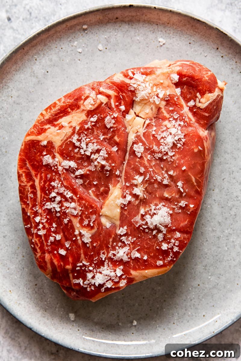 raw ribeye steak sprinkled with flaked salt on grey plate, ready for seasoning