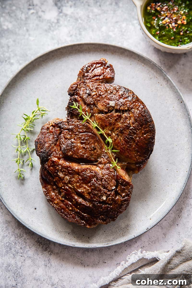 cooked ribeye steak on grey plate and a small dish of herb sauce near it, garnished with fresh herbs