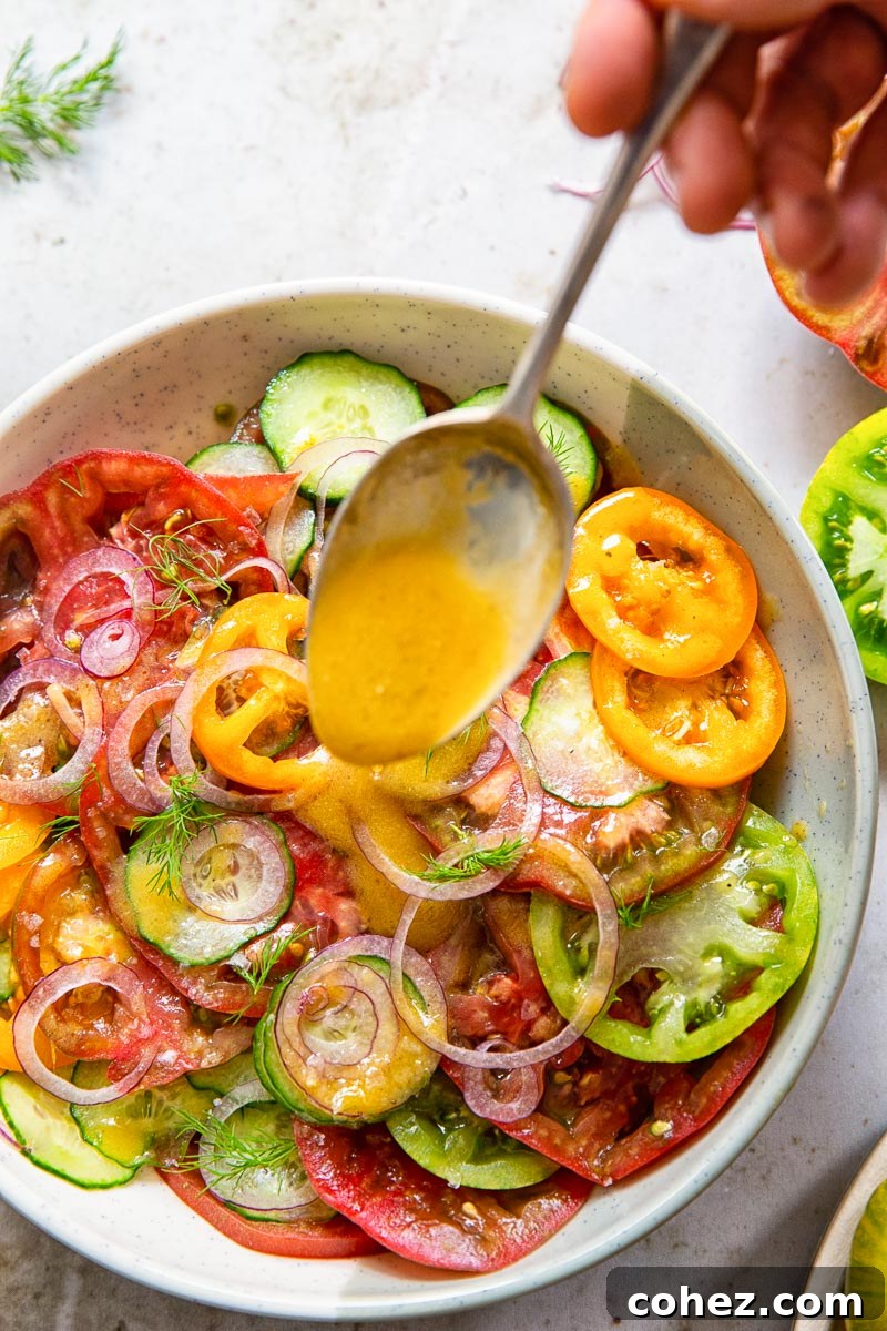 A hand pouring vinaigrette dressing over a bowl of cucumber and tomato salad, highlighting the glistening fresh vegetables