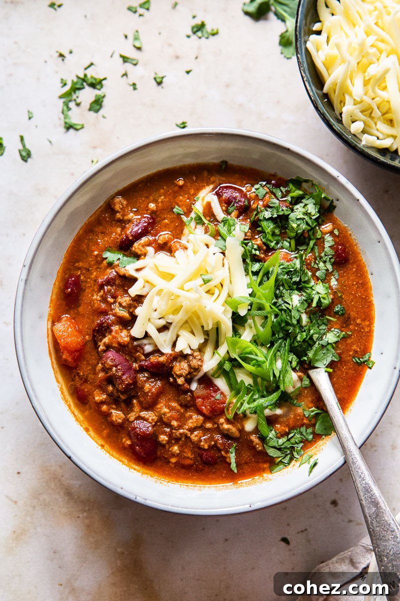 A steaming bowl of chili, generously topped with grated cheese, finely sliced red onions, and fresh cilantro, ready to be enjoyed.