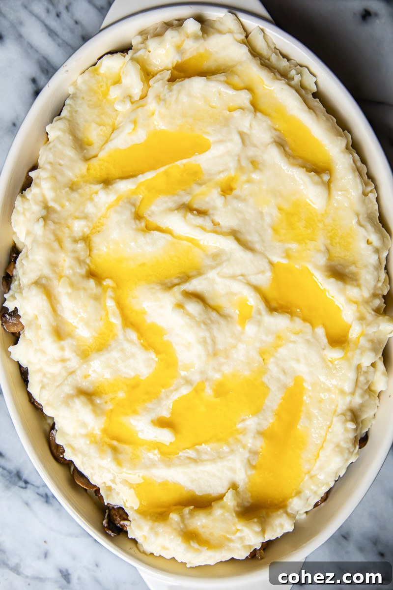 Unbaked mashed potatoes in casserole dish with melted butter drizzled all over, ready for the oven