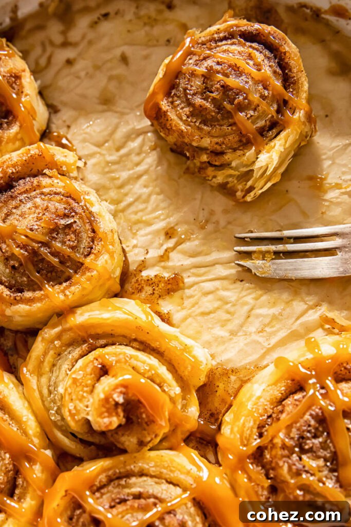 A single puff pastry cinnamon roll is delicately lifted from the baking dish with a fork, showing its warm, flaky layers.
