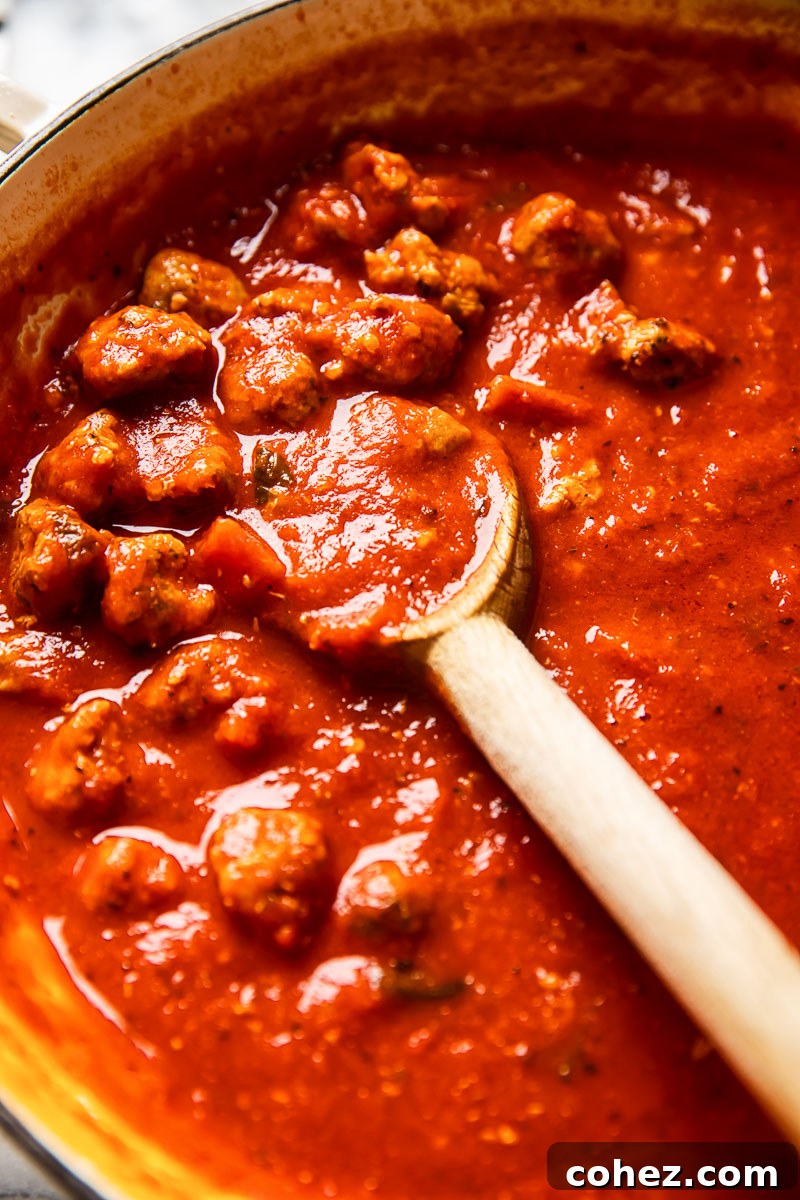 Sausage pasta sauce simmering in a large pan, stirred with a wooden spoon