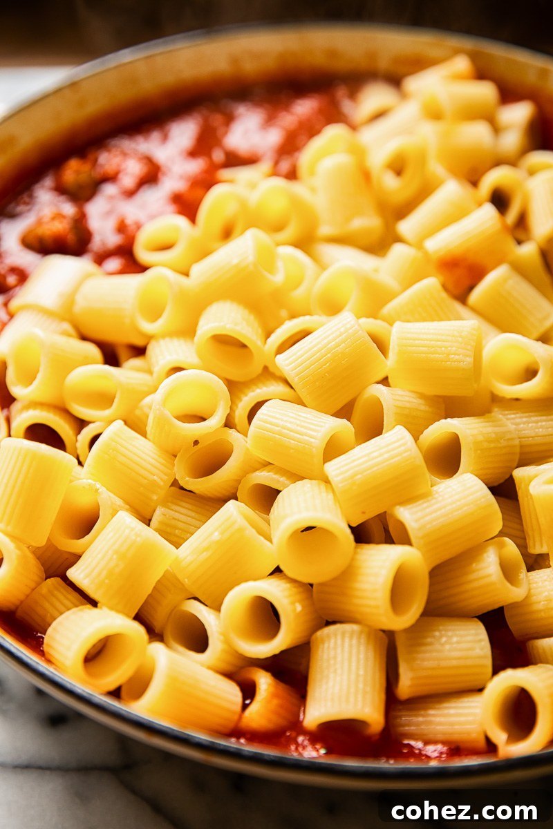 Cooked rigatoni pasta piled over the rich, spicy sausage and tomato sauce in a pan