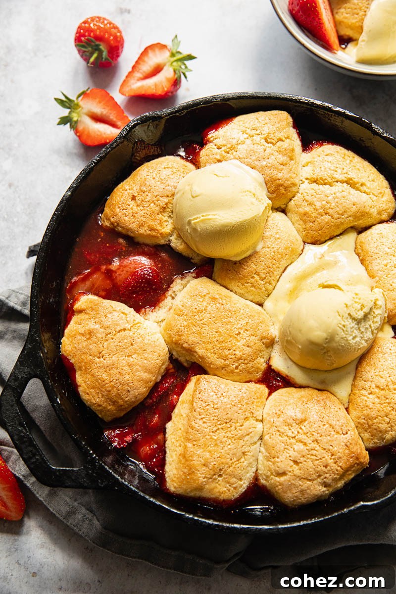 A close-up of a strawberry cobbler in a cast iron skillet, golden brown biscuits topped with a scoop of vanilla ice cream, ready to be served.