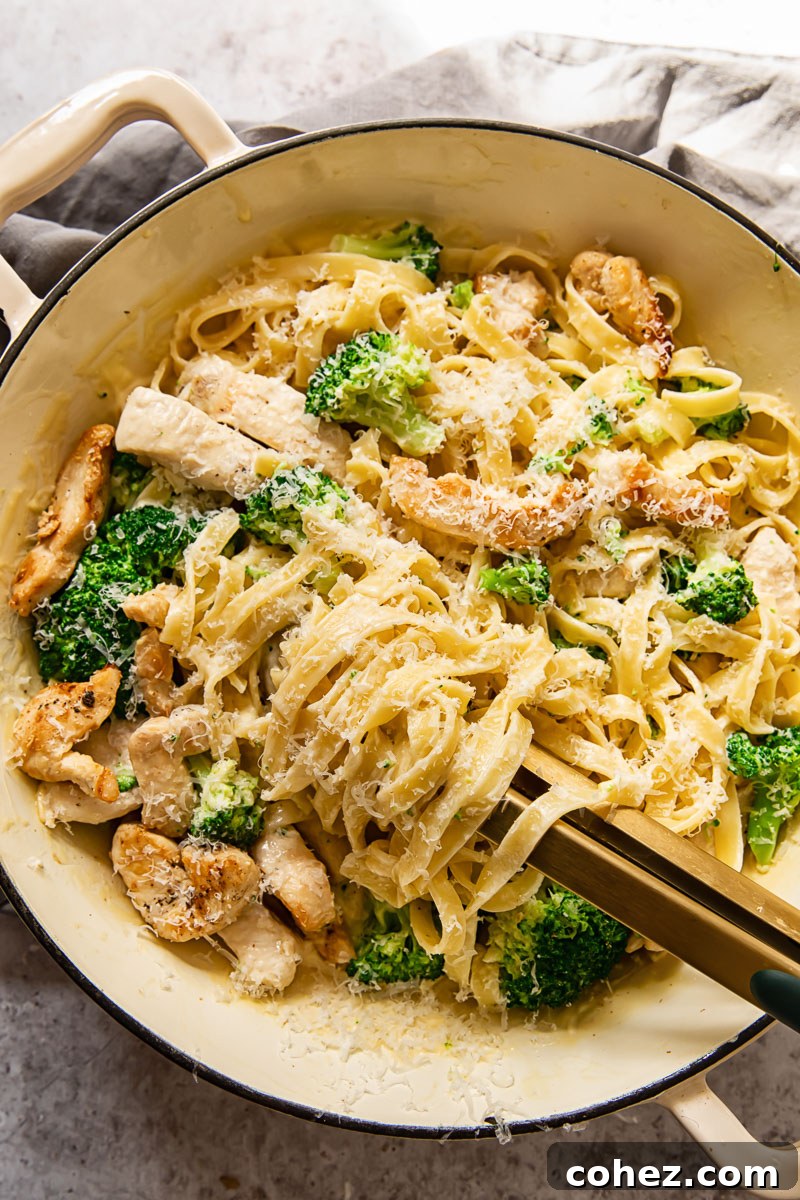 Top-down shot of a large, shallow pan brimming with Chicken Broccoli Alfredo fettuccine, a pair of serving tongs nestled in the pasta, ready to dish out