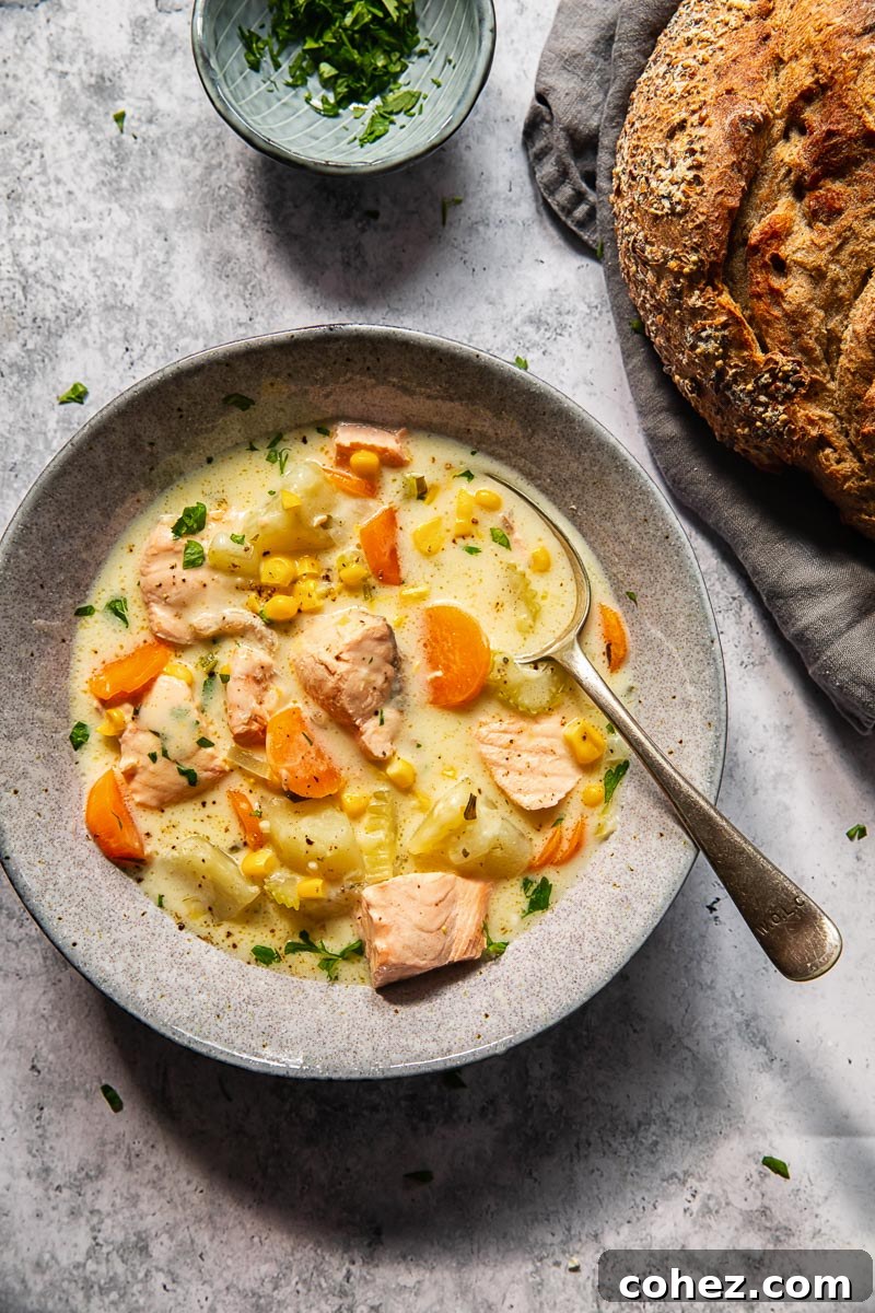 A grey bowl filled with creamy salmon chowder, a spoon dipped in it, with a loaf of rustic bread visible in the background.