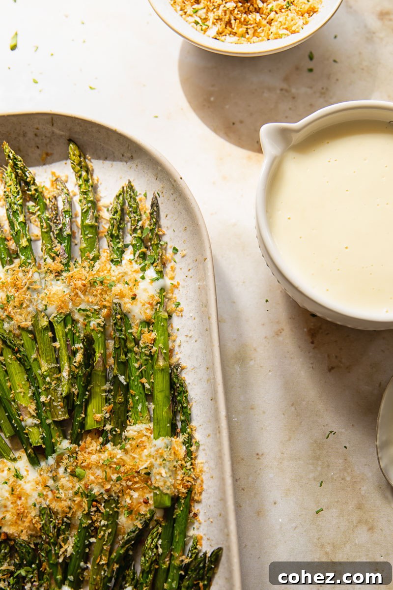 A vibrant platter of roasted asparagus, drizzled with a rich cheese sauce and topped with crunchy breadcrumbs, with a small bowl of extra sauce.