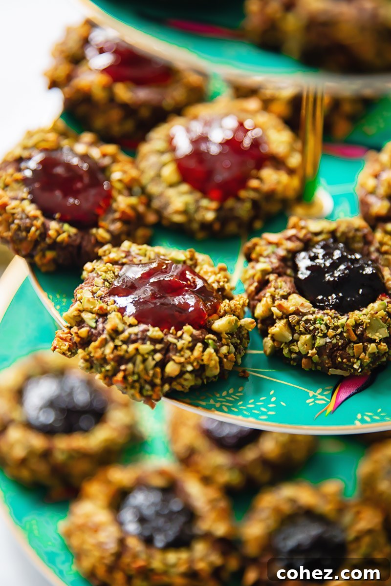 A vibrant close-up shot of several freshly baked chocolate pistachio thumbprint cookies, clearly showing the glistening, jewel-toned jam centers and the intricate, crunchy green pistachio coating.