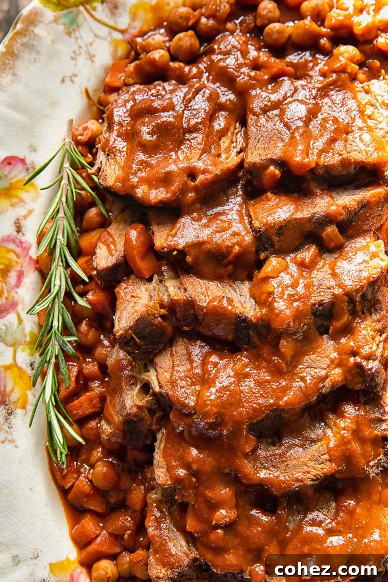 close up of sliced roast beef with a twig of fresh rosemary on the side, in a rich sauce with chickpeas