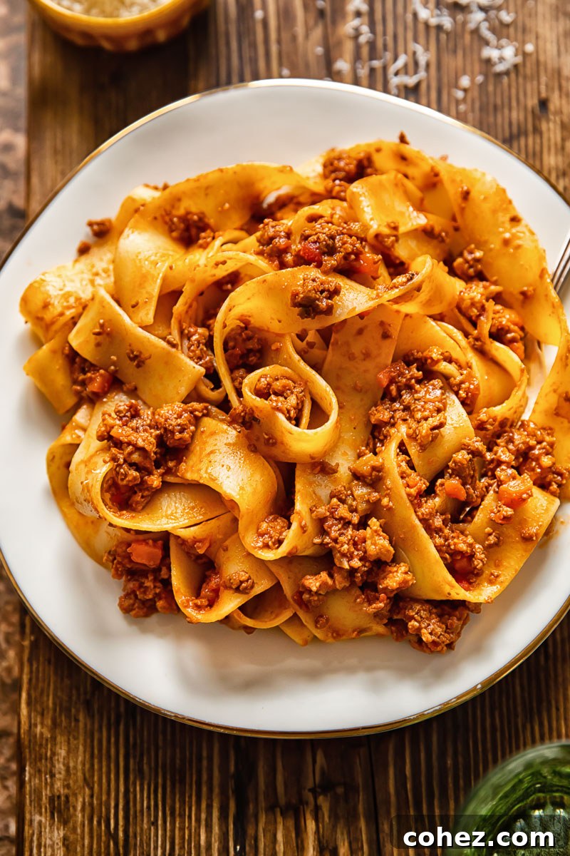 A generous serving of wide pappardelle pasta tossed with a hearty, meaty Tuscan ragu sauce on a pristine white plate, garnished with fresh herbs.