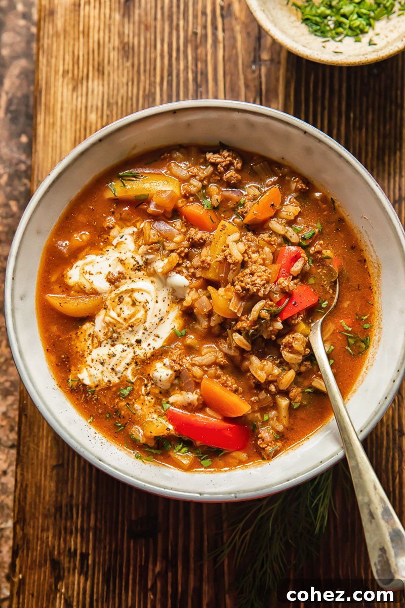 A vibrant bowl of Unstuffed Pepper Soup with a spoon, garnished with fresh herbs, and a small bowl of chopped herbs nearby. The soup features tender bell peppers, savory ground beef, and hearty rice in a rich tomato broth.